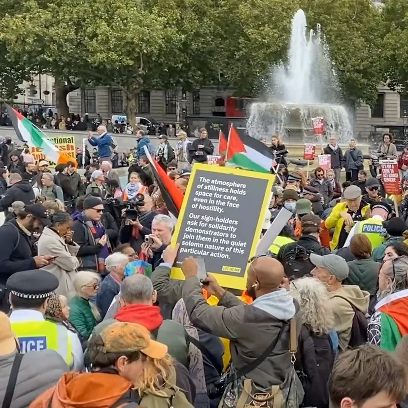 A sign is held aloft at the protest. It reads: "The atmosphere of stillness holds space for care, even in the face of hostility. Our sign-holders ask for solidarity demonstrators to join them in the quiet solemn nature of this aprticular action."